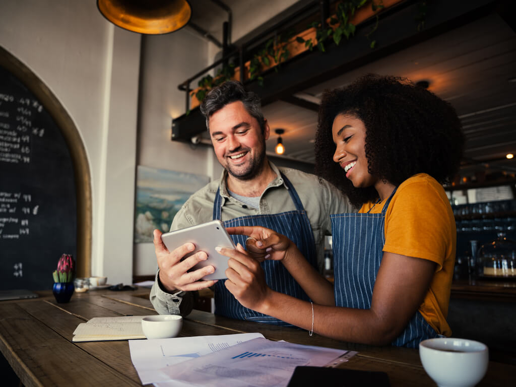 Two employees in a café look at their tablet and are happy about the optimal ERP system